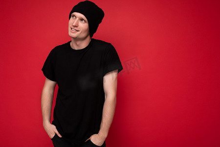 黑色帽子摄影照片_英俊积极的年轻男性站在红色背景墙上孤立的照片，身穿黑色 T 恤作为模型和黑色帽子，看着侧面做梦