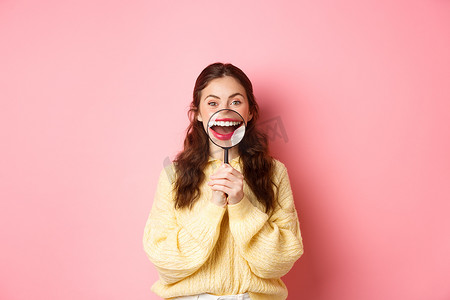 微笑的年轻女性用放大镜展示她完美的美白牙齿微笑，牙科诊所的宣传，口腔医学和医疗保健概念，站在粉红色背景下