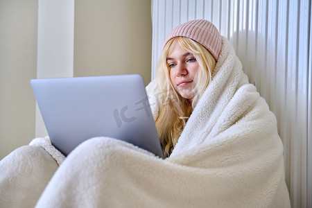 室内取暖摄影照片_冬季寒冷季节冰冻的青少年，用毯子帽取暖，看着笔记本电脑