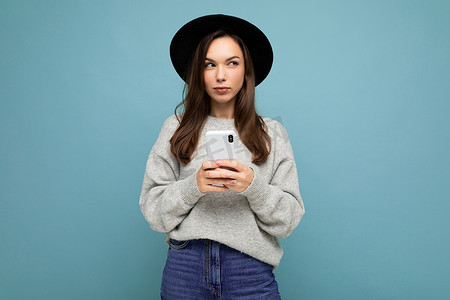 美丽的黑发年轻女性戴着黑色帽子和灰色毛衣，拿着智能手机，看着背景中孤立的侧面发短信