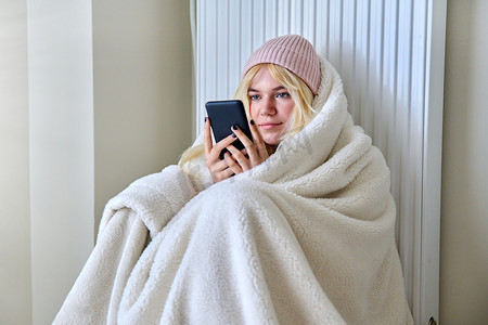 寒冷的季节，戴着帽子的冰冻少年坐在毯子下，坐在带智能手机的暖气片附近