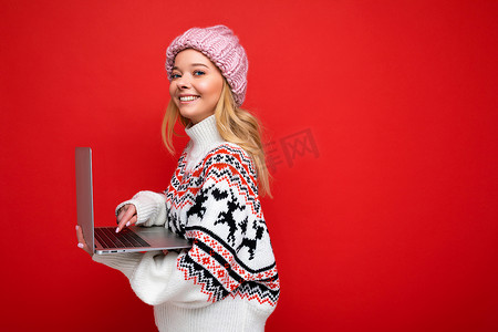 侧面照片中，迷人的微笑快乐漂亮的年轻女子拿着笔记本电脑，隔离在红墙背景上，看着相机，戴着冬帽，穿着针织新年毛衣在键盘上打字