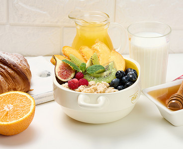 盘子里放着燕麦片和水果，半个成熟的橙子和鲜榨果汁放在透明玻璃瓶中，蜂蜜放在白桌上的碗里。