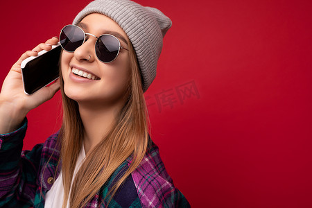 拿着照片摄影照片_美丽微笑的年轻金发女子的特写照片，她穿着时髦的紫色衬衫和休闲的白色 T 恤灰色帽子和太阳镜，隔离在红色背景上，手拿手机，看着旁边的手机