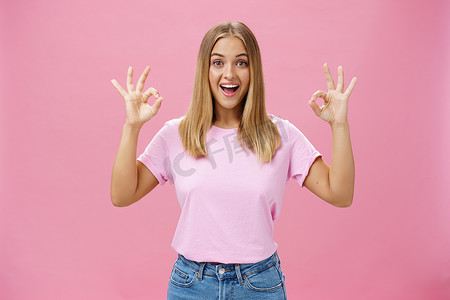 保证手势摄影照片_身着时髦 T 恤和牛仔裤的热情迷人的白人女孩的画像，在粉红色背景下对好消息做出反应，带着愉快的灿烂笑容，表现出好的或确认的手势