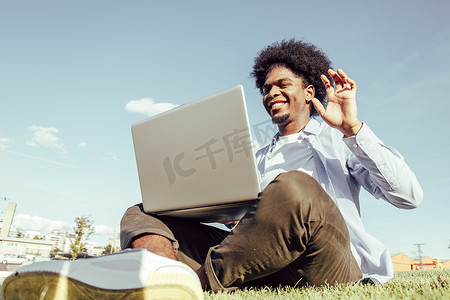 爆炸头摄影照片_留着爆炸头的黑人男子坐在城市公园的草地上使用笔记本电脑。