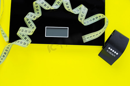 照片上方的黄色背景上放着一个体重秤、一条黑色健身带和一条黄色卷尺。