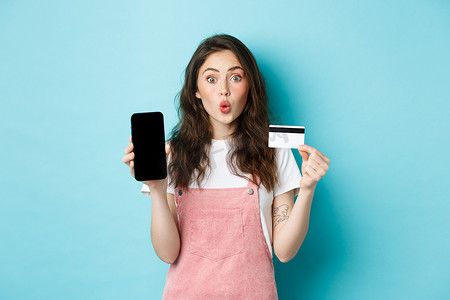 app展示摄影照片_年轻美女的画像展示塑料信用卡和空的智能手机屏幕，看起来既有趣又感兴趣，展示有趣的应用程序，站在蓝色背景上