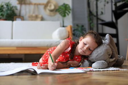 微笑快乐的女孩躺在温暖的地板上，旁边有一只玩具大象，她享受着创造性的活动，用铅笔给相册中的图片上色。