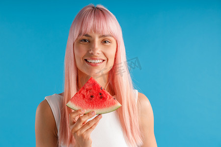 蓝色工作室背景中，一头粉红色头发、拿着西瓜片、对着镜头微笑的美丽年轻女子的画像