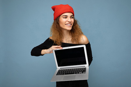 身穿黑色裁剪上衣和红色和橙色抹布的美丽风趣快乐的深色卷发少女，在浅蓝色墙壁背景上被隔离，手持电脑笔记本电脑，屏幕显示器空空如也，面朝侧面