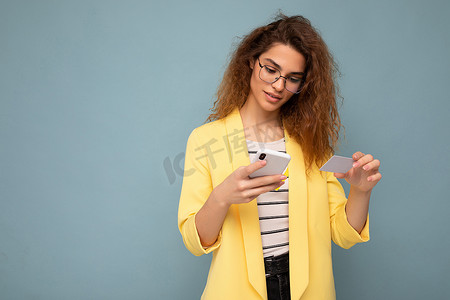好看黄色摄影照片_穿着日常服装的年轻女性在背景中被隔离，手持电话和信用卡通过信用卡支付网上购物费用，看着智能手机屏幕