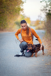 牵狗散步摄影照片_秋季公园里，身穿橙色高领毛衣和牛仔裤的年轻女子牵着狗散步