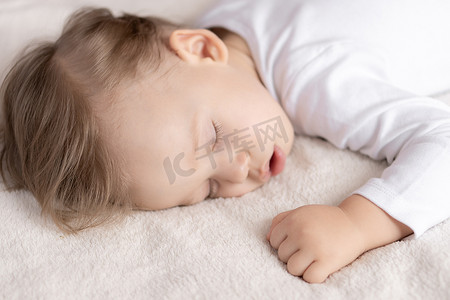 孩子和妈妈睡摄影照片_童年、睡眠、休息、家庭、生活方式概念 — 一个 2 岁可爱小男孩的特写肖像，他身着白色身体，中午睡在米色床上，张着嘴，顶视图和侧视图。