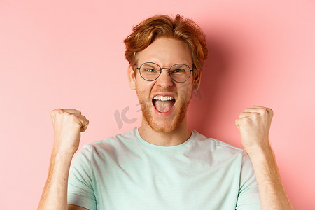 一样高摄影照片_满意的幸运红发男子获胜的特写，高兴地大喊，握拳，庆祝胜利，像冠军一样站在粉红色的背景下