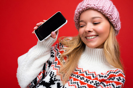 冬季温暖摄影照片_照片中，年轻金发美女微笑着，戴着温暖的针织帽，穿着冬季温暖的毛衣，站在红色背景上，显示智能手机的屏幕为空，可供模拟