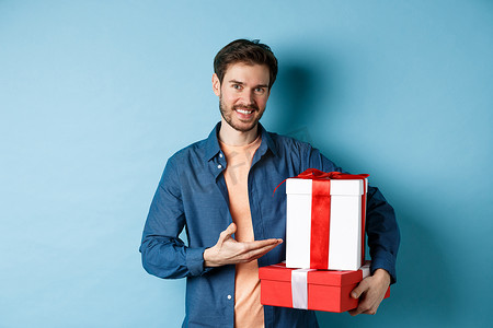 穿着休闲服的年轻人在情人节购买浪漫礼物，指着礼盒微笑，站在蓝色背景上
