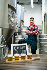 工厂标语牌摄影照片_啤酒取样器托盘上的四杯精酿啤酒和站在背景中的酿酒师