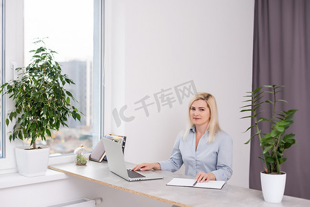 大窗户办公室摄影照片_年轻的金发白人女摄影师坐在笔记本电脑木桌大窗户和灰色墙壁前工作