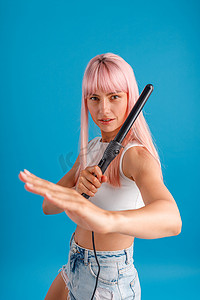圆滑摄影照片_俏皮的粉红色头发的年轻女子看着相机，摆出武术姿势，拿着卷发器，在蓝色工作室背景下摆出孤立的姿势