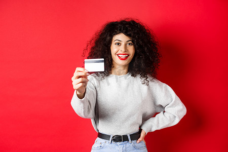 现代女性卷发、伸出手、展示塑料信用卡、推荐银行或购物优惠、红色背景的形象