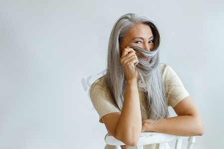 积极的中年亚洲女士坐在工作室的椅子上，用灰白的头发作为面纱
