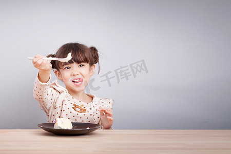 孩子们喜欢吃的概念：快乐的小可爱混血女孩喜欢吃带笑脸的蛋糕，手里拿着勺子的舌头棒，邀请吃。