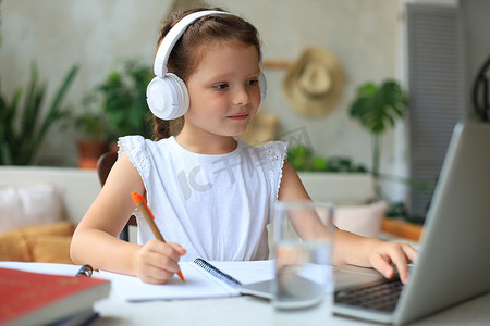 补习学习摄影照片_戴着耳机的微笑小女孩在家中使用笔记本电脑在线手写学习，戴着耳机的可爱快乐小孩子在电脑上上互联网网络课程或课程。