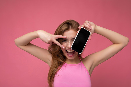 照片中，美丽、积极的年轻金发女子穿着粉红色上衣，在粉红色背景下与世隔绝，手里拿着空的空间，展示手机，手机上有空的显示屏，可以剪下来看着相机，并展示和平姿态
