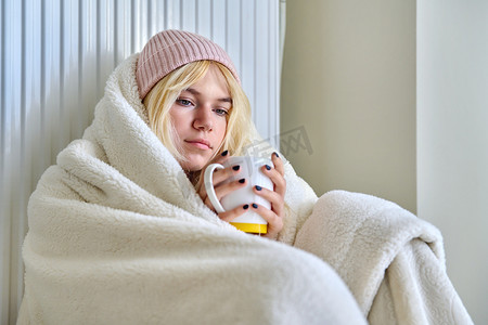 年轻女青少年用毯子、暖气片、热茶取暖