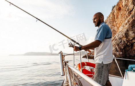 日落时，年轻的非洲裔美国男子拿着钓鱼竿站在帆船上钓鱼
