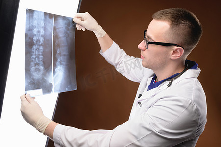 戴着眼镜和医用衣服的年轻医生放射科医生在棕色背景下观察脊柱的管腔 X 射线照片。