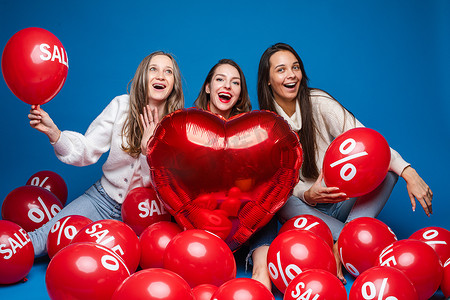 蓝色心空摄影照片_快乐的女性朋友与红色心形气球和空气球合影，蓝色背景上有百分比和销售字母