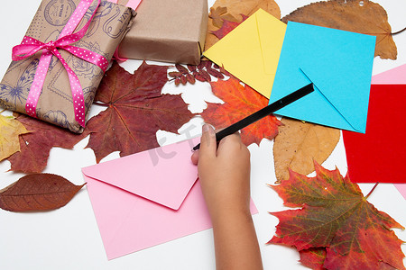 孩子们的手把一张卡片放在信封里，鲜艳的颜色，带漂亮丝带的礼物，节日问候，感恩节，生日，白桌上的秋叶，复制空间顶视图，粉色，黄色，蓝色