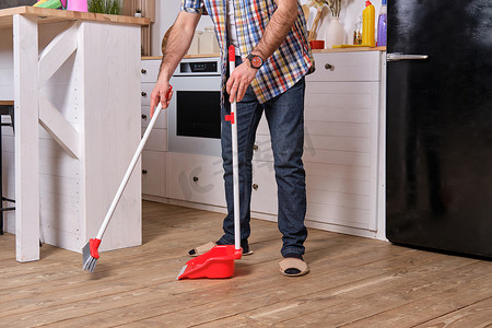 年轻英俊的留着胡子的男人在厨房里，穿着格子衬衫，用铲子和扫帚扫地。