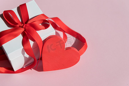 白色礼盒红色丝带摄影照片_情人节的白色礼盒，配有红色蝴蝶结丝带和粉红色背景的红心。生日快乐，婚礼，贺卡，爱情符号。表达感情