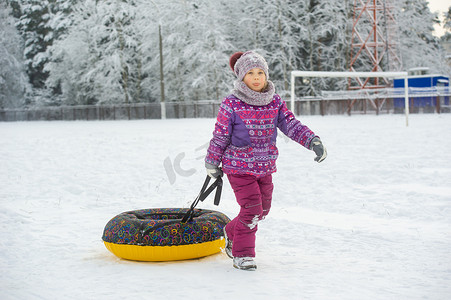 冬天，一个穿着紫色衣服和充气圈的小女孩在白雪覆盖的森林里的街道上行走