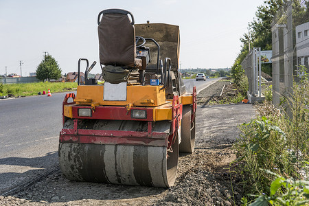 橙色振动压实机站在沥青路附近的地面上，在道路施工和修复沥青路面时与蓝天一起工作。