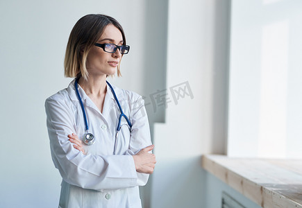 望向窗外摄影照片_身穿医用长袍、戴着眼镜听诊器的女医生望向窗外