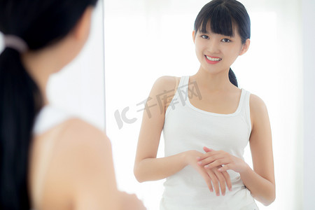 美丽的年轻亚洲女性看着镜子，用手涂抹保湿霜和乳液，以保护皮肤、护肤和化妆品，以实现美容、治疗和身体护理、皮肤护理概念。