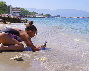 年轻漂亮的女孩坐在海边的海滩上，用手机拍照。夏季。