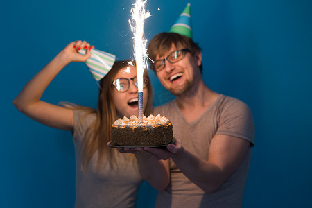 快乐的年轻夫妇迷人的男人和戴着纸帽子的可爱的女孩做出愚蠢的表情，手里拿着一个站在蓝色背景上的生日蛋糕。