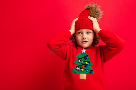圣诞毛衣摄影照片_穿着红色圣诞毛衣的酷少年在红墙、温暖的帽子和带圣诞树的毛衣的背景下闲逛