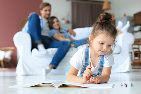 微笑幸福的家庭坐在客厅的沙发上放松地看着小女儿用彩色铅笔在相册里画画。