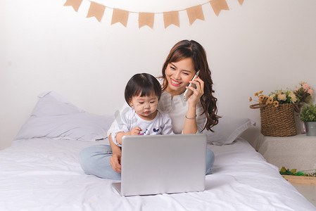 女士西装摄影照片_穿着经典西装的亚洲女士正在用手机交谈，并与她的女婴在家里使用笔记本电脑。