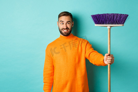 打扫房子摄影照片_男朋友对扫帚感到困惑，强迫打扫房子，做家务，在浅蓝色背景下站着不高兴