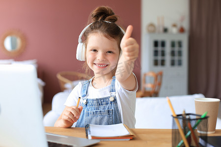 快乐的小女孩在家使用笔记本电脑在线学习，微笑的小孩子竖起大拇指推荐课程或课程。