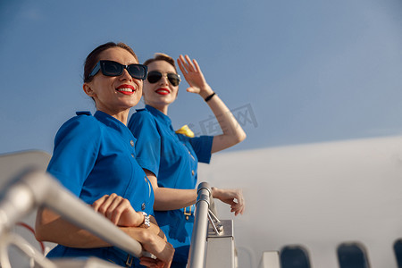 阳光明媚的日子里，两位身穿蓝色制服、戴着墨镜、开朗的空姐微笑着站在楼梯上的肖像