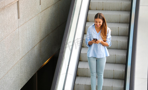 电梯乘坐摄影照片_年轻的女商人在乘坐自动扶梯下降时用智能手机发送消息。