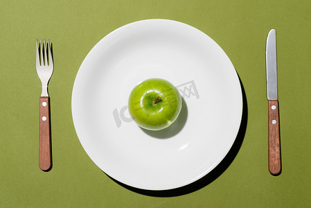白盘上青苹果的顶部视图，绿色背景上有刀叉